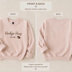May include: A light pink crewneck sweatshirt featuring the embroidered text Hedge-Hug CLUB with a small hedgehog graphic on the front. The garment has long sleeves with ribbed cuffs and a ribbed hem. The back of the sweatshirt is plain.