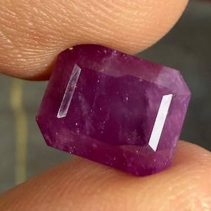 Puede incluir: Una piedra preciosa rectangular facetada con un tono magenta púrpura intenso. La piedra presenta una superficie clara y pulida con inclusiones internas visibles y bordes afilados y angulares que captan la luz de forma vibrante.