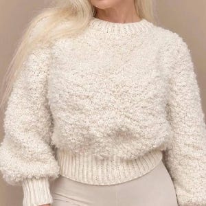 Puede incluir: Un suéter de manga larga color crema con un patrón de tejido texturizado, difuso y similar a las palomitas de maíz. La prenda tiene un cuello redondo, un dobladillo de punto acanalado y puños acanalados en las mangas.