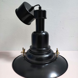 Puede incluir: Lámpara de escritorio de metal negro con base redondeada y pantalla cónica. La lámpara tiene un brazo flexible y un cabezal pequeño y ajustable. La lámpara mide aproximadamente 30 cm de altura y tiene detalles en latón.