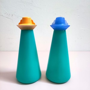 May include: Two teal-colored, cone-shaped salt and pepper shakers. One has an orange top, the other a blue top. The tops are shaped like small hats. The shakers are on a white surface.