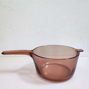 Puede incluir: Un cazo de cristal translúcido de color ámbar con un recipiente redondo, un mango largo que se extiende por un lado y una pequeña asa en forma de lengüeta en el lado opuesto. El utensilio tiene un acabado liso y un tono marrón uniforme.