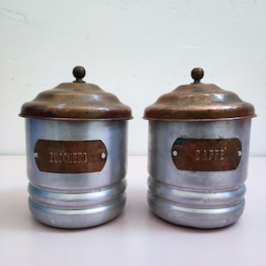 Op de afbeelding: Twee vintage metalen canisters met koperen deksels en kleine knoppen. Op een canister staat "ZUCCHERO" en op de andere "CAFFE". De canisters hebben een zilveren behuizing met een geribbeld ontwerp en zijn waarschijnlijk voor het bewaren van koffie en suiker.