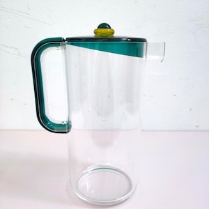 May include: A clear glass pitcher with a green handle and lid. The lid has a yellow and green knob. The pitcher has a spout and a capacity of approximately 1 liter. The design is modern and minimalist, suitable for serving beverages.