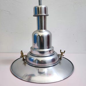 Puede incluir: Una lámpara colgante plateada pulida con una pantalla circular y un cuerpo cilíndrico. La luz tiene un acabado metálico y está suspendida por un cable negro. El accesorio tiene detalles de latón.