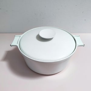 May include: A round, white enamelled cooking pot with a matching lid and two integrated side handles. The pot features a smooth, solid white finish throughout, including the circular knob on the centre of the lid and the wide, flat side handles.