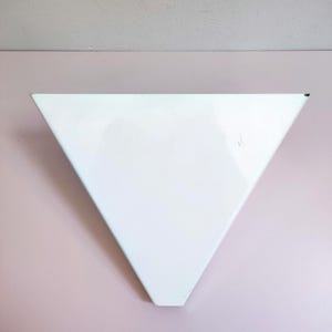 May include: A white, triangular wall shelf. The shelf is made of metal and has a smooth, glossy finish. The shelf is mounted on a pink wall.