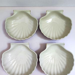 Op de afbeelding: Een set van vier gebroken witte keramische schaaltjes in de vorm van sint-jakobsschelpen. Elk schaaltje heeft een glanzende afwerking met reliëf in de vorm van radiale ribbels die vanuit het scharnier naar de geschulpte rand lopen.