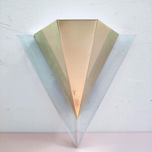 May include: Triangular wall sconce with a geometric design. The sconce has a central section in a warm gold colour, with gold and green sections on either side. The lower part is a clear, triangular shape. It is mounted on a white wall.