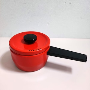 Puede incluir: Una pequeña cacerola roja con asa y tapa negras. La tapa tiene un pomo negro y varios agujeros pequeños. La cacerola está sobre una superficie rosa claro.