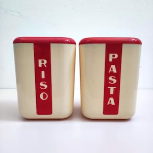 Könnte beinhalten: Zwei Vintage-Küchenbehälter. Die Behälter sind cremefarben mit roten Akzenten. Auf einem Behälter steht "RISO" in Rot, auf dem anderen "PASTA" in Rot. Die Deckel sind rot.