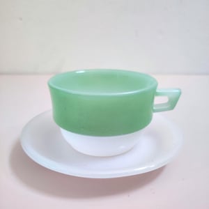 Op de afbeelding: Een vintage kop en schotel set. De beker is tweekleurig, met een lichtgroene bovenkant en een witte basis. De schotel is wit. De beker heeft een klein, rechthoekig handvat. De set is waarschijnlijk gemaakt van glas of keramiek.