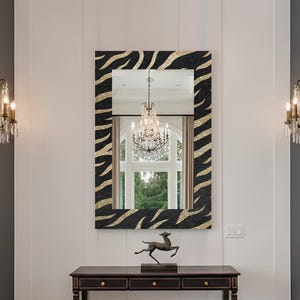 Può includere: Uno specchio rettangolare con una cornice con stampa zebrata nera e oro è appeso sopra una consolle in legno scuro. Una panca in velluto verde con gambe dorate è a sinistra. Lo specchio riflette un lampadario e una finestra. Due applique dorate sono montate a parete.