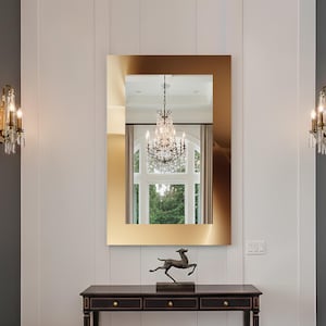 Puede incluir: Un espejo rectangular con marco dorado refleja una lámpara de araña y una ventana. Debajo, una consola de madera oscura con tres cajones sostiene una escultura de ciervo de bronce. Un banco de terciopelo verde y apliques de pared completan la elegante entrada.