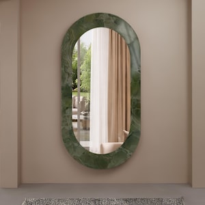 Peut inclure: Un miroir mural de forme ovale doté d'un cadre large et épais avec une finition en marbre vert poli. La surface en verre réfléchissant offre une vue claire sur une pièce intérieure avec des rideaux beiges et un aperçu d'un jardin extérieur.