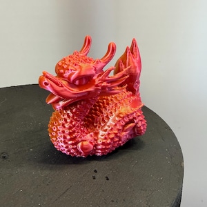 Puede incluir: Figura de dragón roja impresa en 3D. El dragón tiene escamas detalladas, cuernos y una cola enroscada. La figura está sobre una superficie negra. El color del dragón cambia de rojo a naranja.