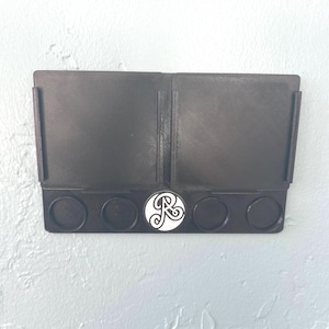 Può includere: Organizzatore da parete nero con due scomparti rettangolari e quattro supporti circolari. Un emblema bianco e nero è centrato in basso. L'organizzatore è montato su una parete azzurra.