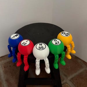 May include: Five spherical bingo ball figures with articulated legs in blue, red, white, green, and yellow. Each ball features a letter and number: B7, I22, N35, G47, and O69. The figures are arranged in a row, each with a round body and jointed limbs.