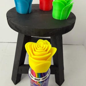 Puede incluir: Un pequeño taburete de madera negro exhibe tres esculturas de rosas impresas en 3D en azul, rojo y verde. Una escultura de rosa amarilla se coloca sobre una botella morada y arcoíris con la palabra "VIOLET" impresa.