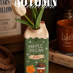 Peut inclure: Un vase en céramique en forme de brique de lait, de couleur vert sauge et crème, portant l'inscription "MAPLE PECAN latte" et des illustrations d'une tasse de café, d'une feuille d'érable et de noix de pécan. Trois tulipes blanches sont insérées.
