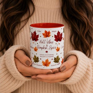 May include: A white ceramic mug with a bright red interior, featuring a colorful pattern of autumn leaves in shades of red, orange, and green. The text reads Fall Vibes & Pumpkin Spice, Leaves Are Falling, Autumn Is Calling, Cider, Maple, and more.