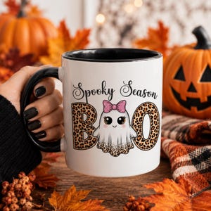 May include: A white ceramic mug with a black handle and interior rim. The front features the text Spooky Season in black script above a graphic of a white ghost with a pink glitter bow and leopard print accents, flanked by the letters B and O.