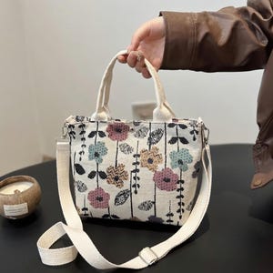 Puede incluir: Un bolso tote pequeño con estampado floral sobre fondo crema. El diseño incluye flores rosas, azules y amarillas con tallos negros. Tiene asas color crema y una correa de hombro desmontable.