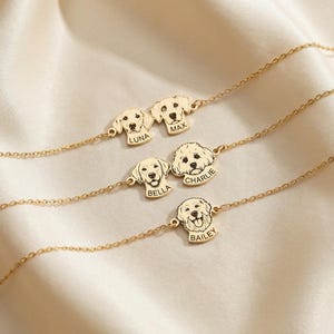 May include: Gold-colored dog-themed necklaces with personalized dog faces and names: Luna, Max, Bella, Charlie, and Bailey. The necklaces are displayed on a cream-colored satin surface.