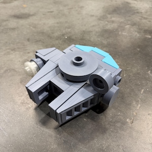 Könnte beinhalten: Ein graues und blaues Modellraumschiff, das aus ineinandergreifenden Blöcken besteht. Das Modell hat eine zentrale kreisförmige Komponente, zwei Seitenflügel und einen hellblauen Abschnitt oben. Ein klares, zylindrisches Teil ist an der Seite befestigt.