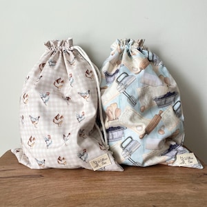 May include: Two drawstring bags. One bag features a beige gingham pattern with chicken illustrations. The other bag has a light blue background with baking-themed illustrations, including mixers, eggs, and rolling pins. Both bags have a white drawstring closure.