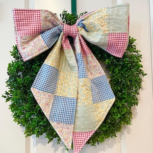 以下が含まれることがあります： 鮮やかな緑色のベースに、大きなパッチワークのリボンが飾られた装飾的なリース。リボンは、ピンク、青、黄色のギンガム、花柄、水玉模様を組み合わせています。緑のリボンは、リースを吊るすために使用されています。