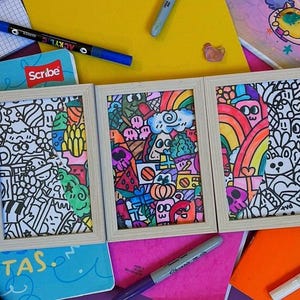 May include: Three framed doodle art pieces with colourful illustrations of various objects, including rainbows, skulls, and food items. The frames are light-coloured wood, and the artwork is set against a backdrop of notebooks and markers.
