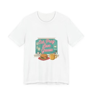 May include: White t-shirt featuring a diner-style graphic with the text "Eat Pray Love Grease." The design includes a burger, a cup of coffee, and decorative elements. The shirt is a casual wear item.