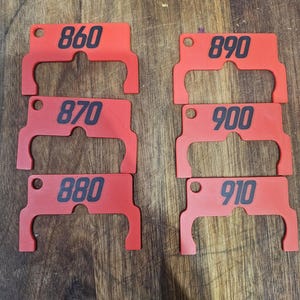 以下が含まれることがあります： 黒い数字が印刷された6つの赤い長方形のタグ。数字は860、870、880、890、900、910です。各タグには左側に小さな穴があり、2つの湾曲した切り欠きを持つ独特の形状をしています。