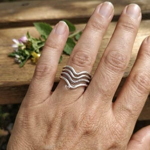 May include: A silver ring set on a finger, featuring a wavy design with a rope-like texture. The ring is composed of multiple bands, creating a layered effect. The background includes a wooden surface and a few flowers.