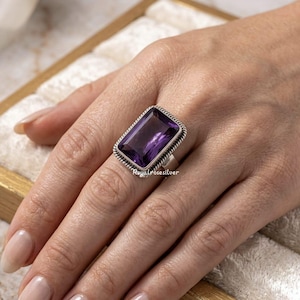 May include: A rectangular amethyst gemstone ring. The ring features a large, deep purple gemstone set in a silver band with a textured border. The ring is displayed on a person's finger, showcasing its size and design.