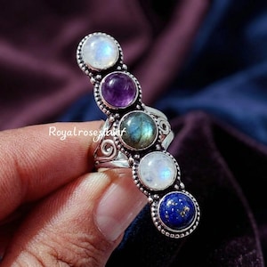 Pode incluir: Um anel de prata com cinco pedras preciosas coloridas: uma pedra da lua branca, uma ametista roxa, uma labradorita verde, outra pedra da lua branca e um lápis-lazúli azul. O anel tem um design ornamentado com o texto "Royalrosesilver".