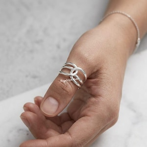 May include: A silver ring with an unusual, interwoven design. The ring has multiple bands that loop and cross over each other, creating an open, geometric shape. The ring is worn on a finger, showcasing its intricate details and polished finish. The ring is a piece of jewellery.