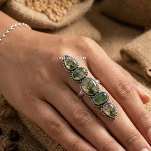 May include: A long silver ring featuring five oval and teardrop-shaped moss agate stones with green inclusions, set in a twisted metal frame. The text royal rose silver is visible on the ring. The design covers a significant portion of the finger.