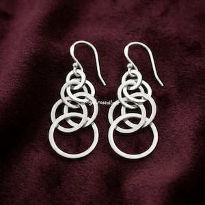 Puede incluir: Un par de pendientes plateados con un diseño de círculos en cascada. Cada pendiente presenta múltiples anillos interconectados de varios tamaños, creando un efecto en capas. Los pendientes cuelgan de ganchos curvos, sobre un fondo burdeos oscuro.