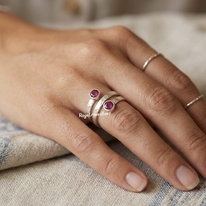 Puede incluir: Un anillo de plata con textura martillada, con dos gemas redondas de color rosa intenso. El anillo se enrolla alrededor del dedo, con las gemas en la parte superior. El nombre de la marca "Royalrosesilver" es visible.