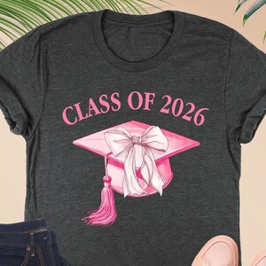 May include: Dark grey heather t-shirt with the text "CLASS OF 2026" and a pink graduation cap graphic. The image also includes a pair of dark wash jeans, a small pink wallet, and a pair of pink sneakers.