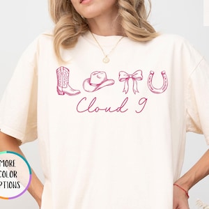 May include: Ivory-colored t-shirt with a pink graphic of a cowboy boot, hat, bow, and horseshoe, with the words "Cloud 9" in cursive. The shirt has short sleeves and a relaxed fit.
