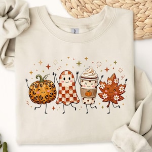May include: A sand-colored crewneck sweatshirt featuring a graphic print of four cartoon characters: a leopard-print pumpkin, a checkered ghost, a coffee cup with whipped cream, and an autumn leaf, all with smiling faces and thin limbs.