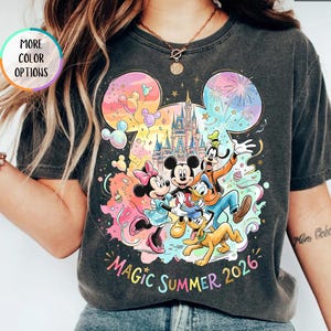 K&ouml;nnte beinhalten: Dunkelgraues T-Shirt mit einem farbenfrohen Aufdruck von Mickey Mouse, Minnie Mouse, Donald Duck, Goofy und Pluto vor einem Schloss. Der Text "Magic Summer 2026" ist darunter gedruckt. "More Color Options" steht in einem Kreis links.