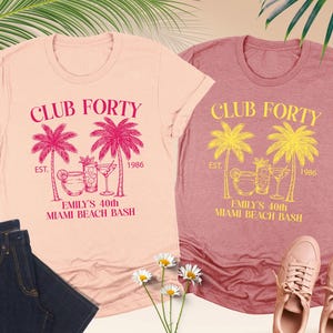 May include: Two heather t-shirts with the text "CLUB FORTY" and "EMILY'S 40th MIAMI BEACH BASH". One shirt is peach with pink graphics, the other is mauve with yellow graphics. The image also shows a pair of jeans, sneakers, and flowers.