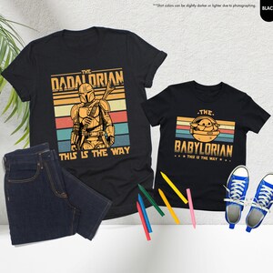 以下が含まれることがあります： スターウォーズをテーマにしたグラフィックがプリントされた黒いTシャツ2枚。「The Dadalorian」とマンダロリアン、「The Babylorian」とグローグーが描かれています。両方に「This is the Way」の文字。青いジーンズと青いスニーカーも写っています。