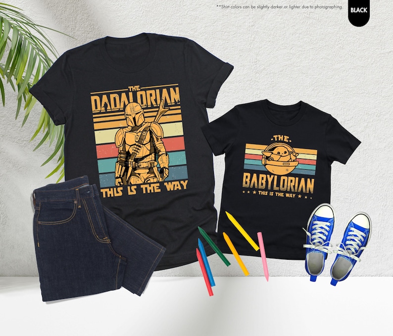 以下が含まれることがあります： スターウォーズをテーマにしたグラフィックがプリントされた黒いTシャツ2枚。「The Dadalorian」とマンダロリアン、「The Babylorian」とグローグーが描かれています。両方に「This is the Way」の文字。青いジーンズと青いスニーカーも写っています。