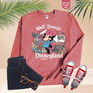 Pu&ograve; includere: Felpa rosso scarlatto melange con grafica di Minnie Mouse e la scritta "Walt Disney's Disneyland Est.1955". L'immagine include anche jeans blu scuro, scarpe da ginnastica rosse e occhiali.