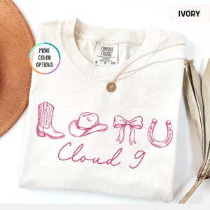 May include: Ivory-colored t-shirt with a pink graphic of a cowboy boot, hat, bow, and horseshoe, with the words "Cloud 9" in cursive. The shirt has a crew neck and short sleeves. A gold necklace is draped on the shirt.